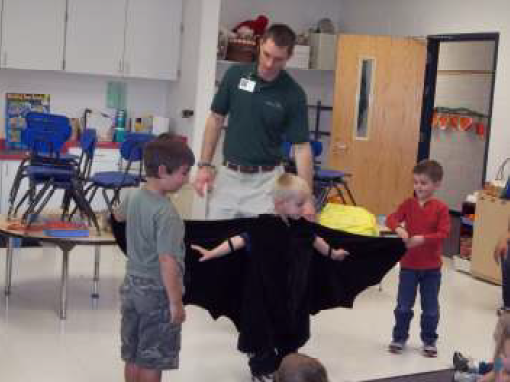 Young boy is dressed like a bat with arms spread out for flying. Two boys watch and adult is talking behind the boys.
