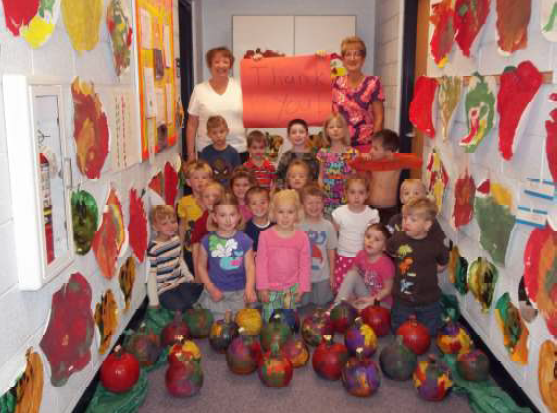 Students in hallway kneeling with painted pumpkins in front of them. Hallway has pictures of pumpkins and other fall items.