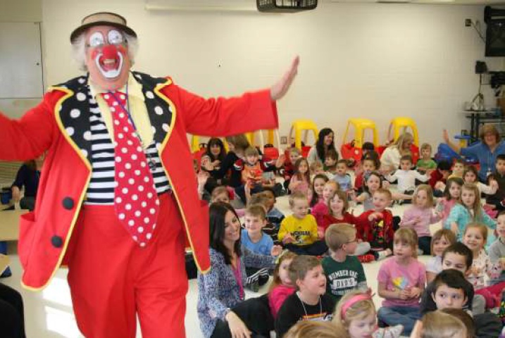 Clown Dressed in bright red walking through assembly of students and teachers (sitting on floor)